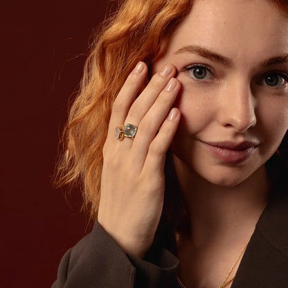 A close-up lifestyle shot of a woman with red hair wearing a gold ring with a blue topaz stone, posing with her hand near her face against a dark reddish-brown background.