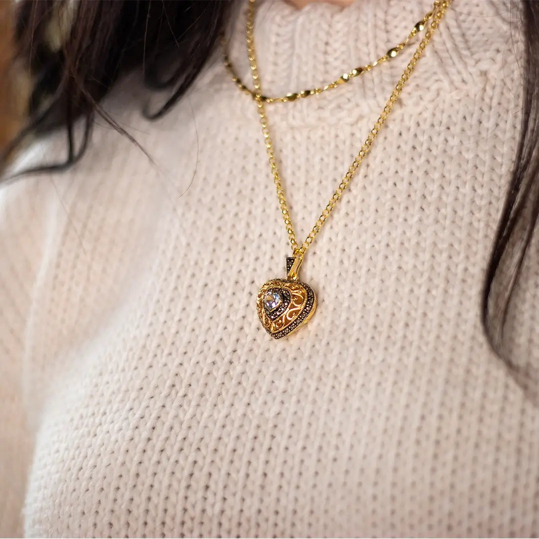 Close-up of a gold heart-shaped locket with intricate vintage detailing and a blue topaz gemstone centre, worn over a cream knit sweater.