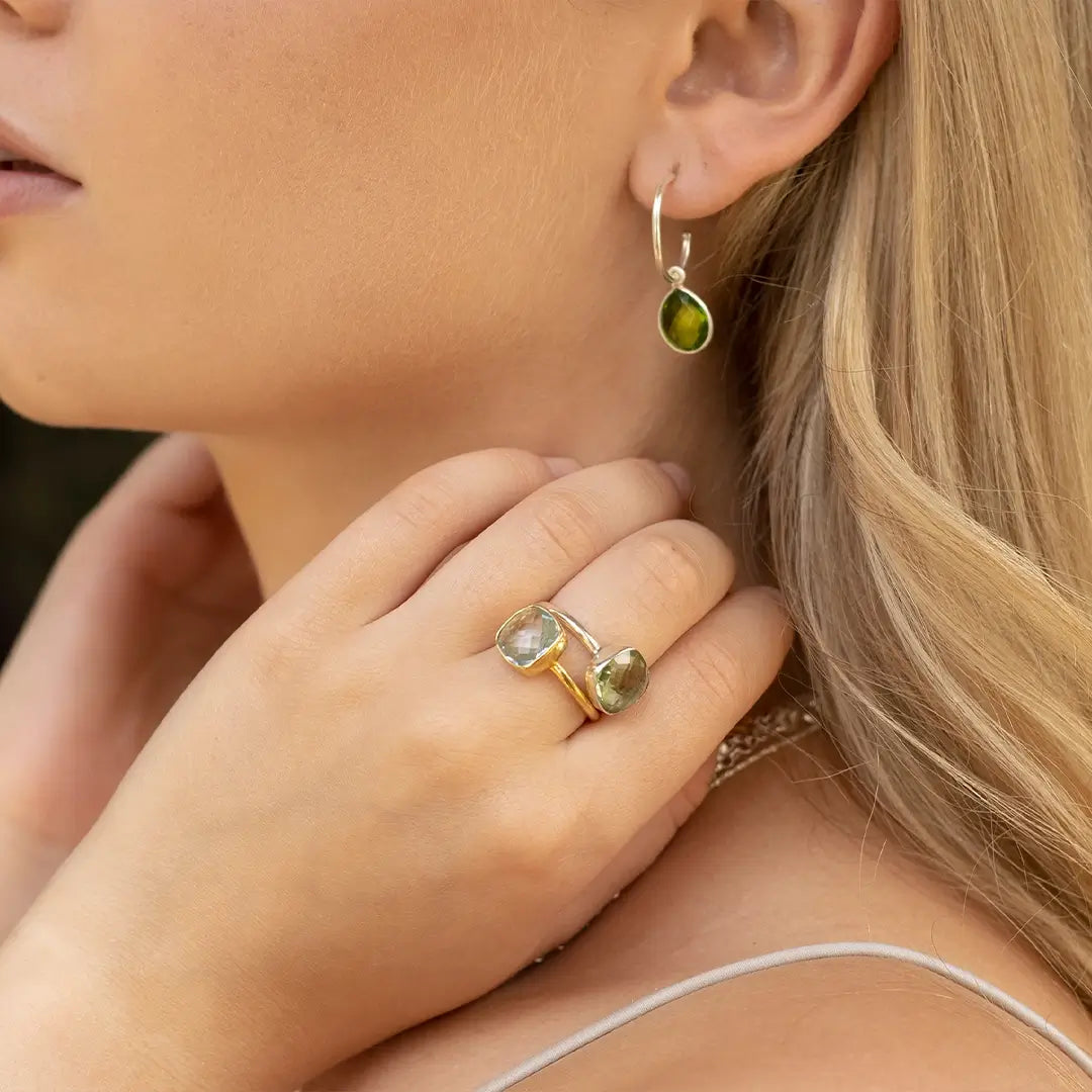 Close-up of a woman wearing green gemstone drop hoop earrings and a gold open ring set with two faceted gemstones, styled against blonde hair and natural skin tones.
