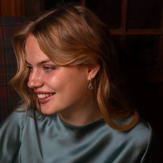 Woman smiling and looking to the side, wearing a teal satin top and silver hoop earrings with teardrop-shaped blue topaz gemstone.