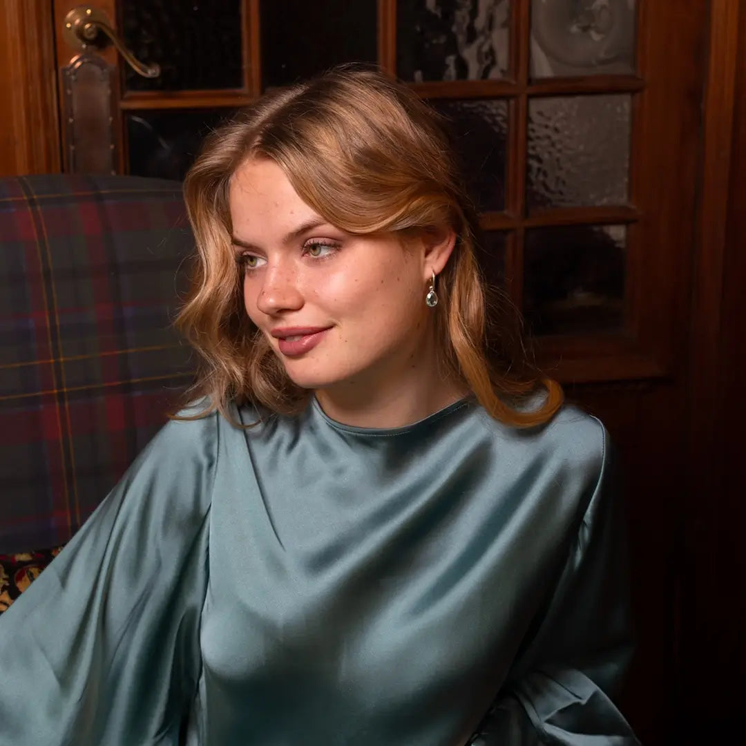 Woman wearing a teal satin blouse and silver hoop earrings with teardrop-shaped blue topaz gemstones, sitting indoors and looking to the side.