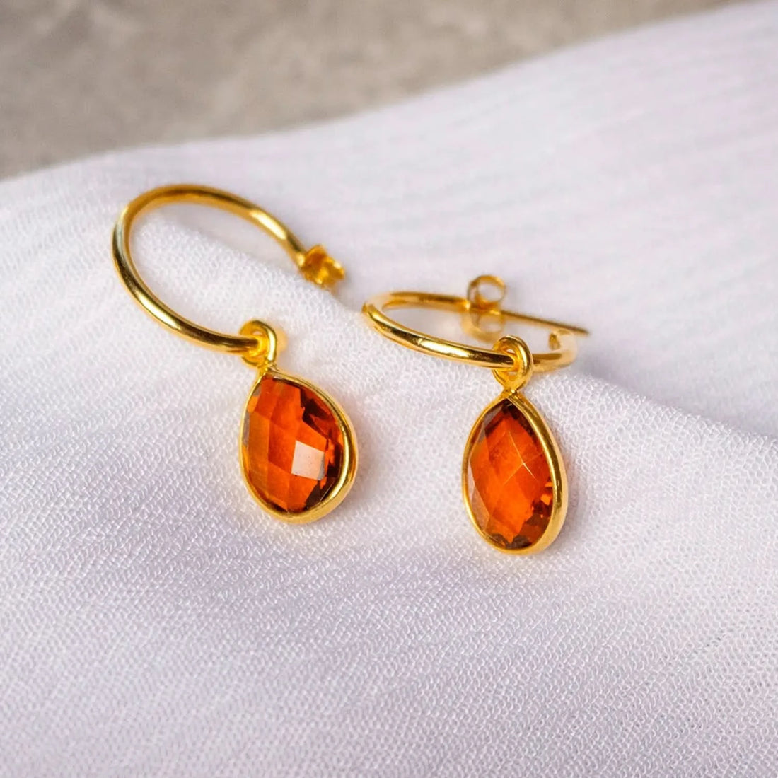 a pair of yellow gold hoop earrings with teardrop shaped carnelian gemstone lying on their side on a white piece of cloth. 