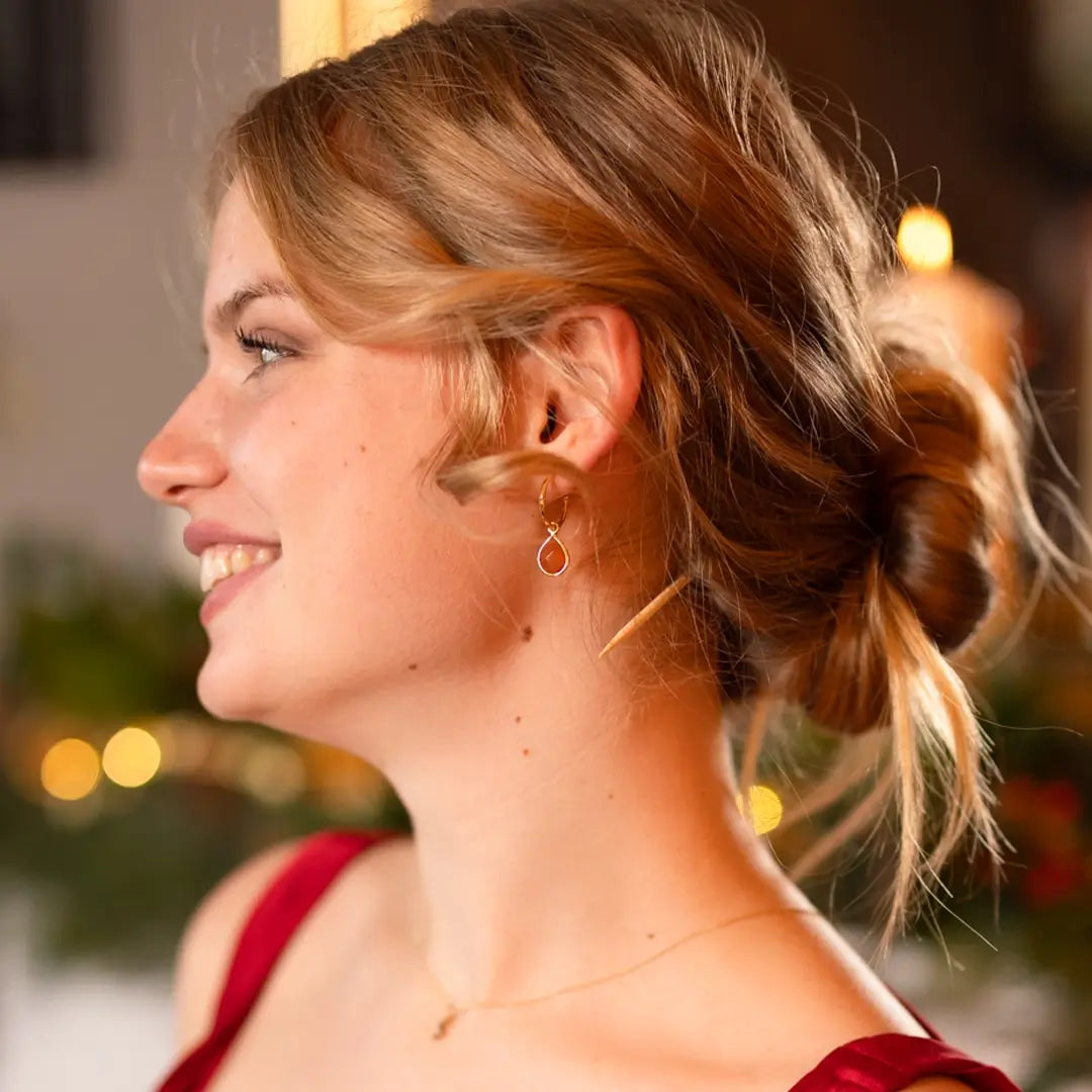 Smiling woman wearing a red dress and gold hoop earrings with carnelian gemstone drops, styled with a low bun and warm background lights.
