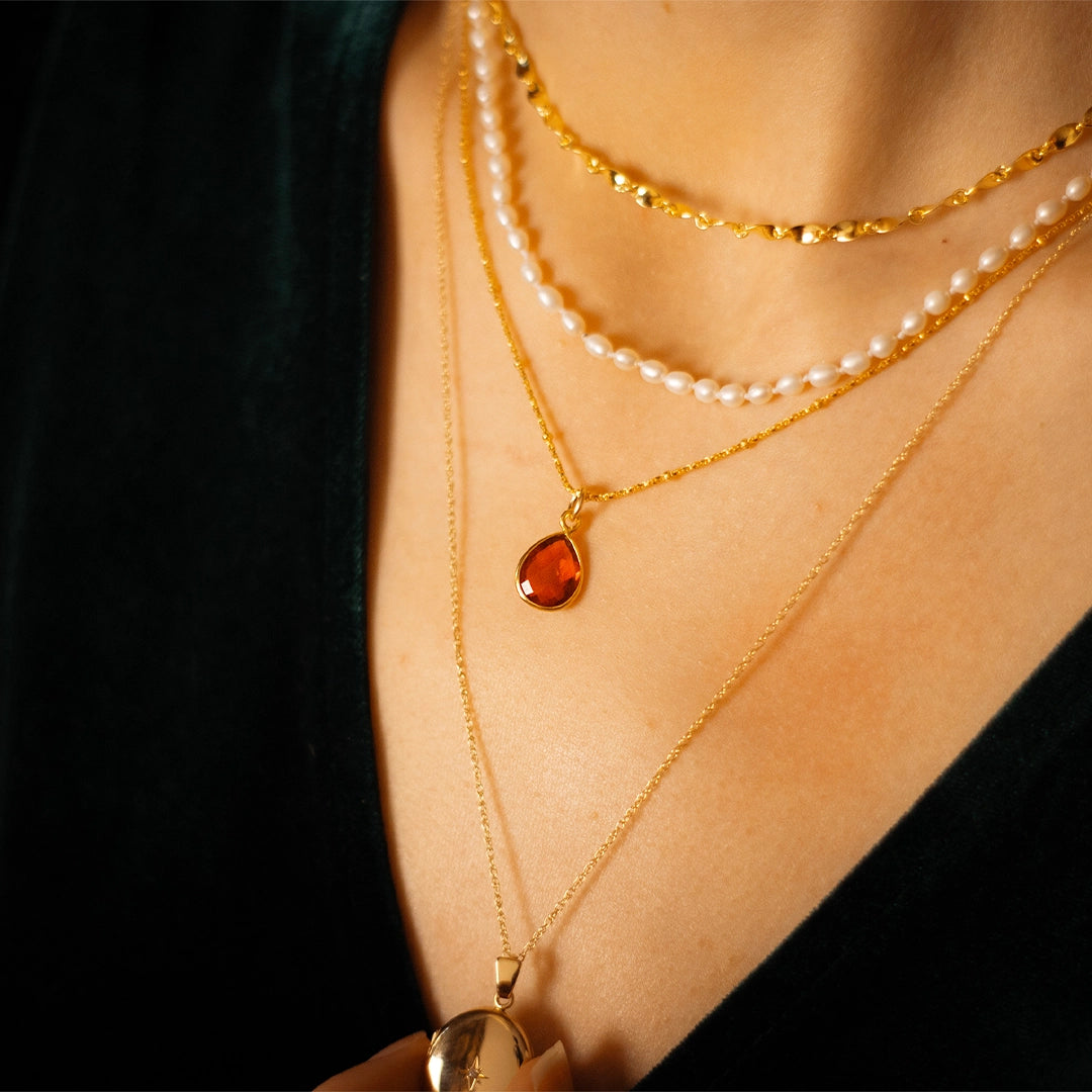 Layered gold necklaces worn on the neck, featuring a teardrop-shaped carnelian gemstone pendant, a delicate pearl strand, and fine gold chains styled against a black top.