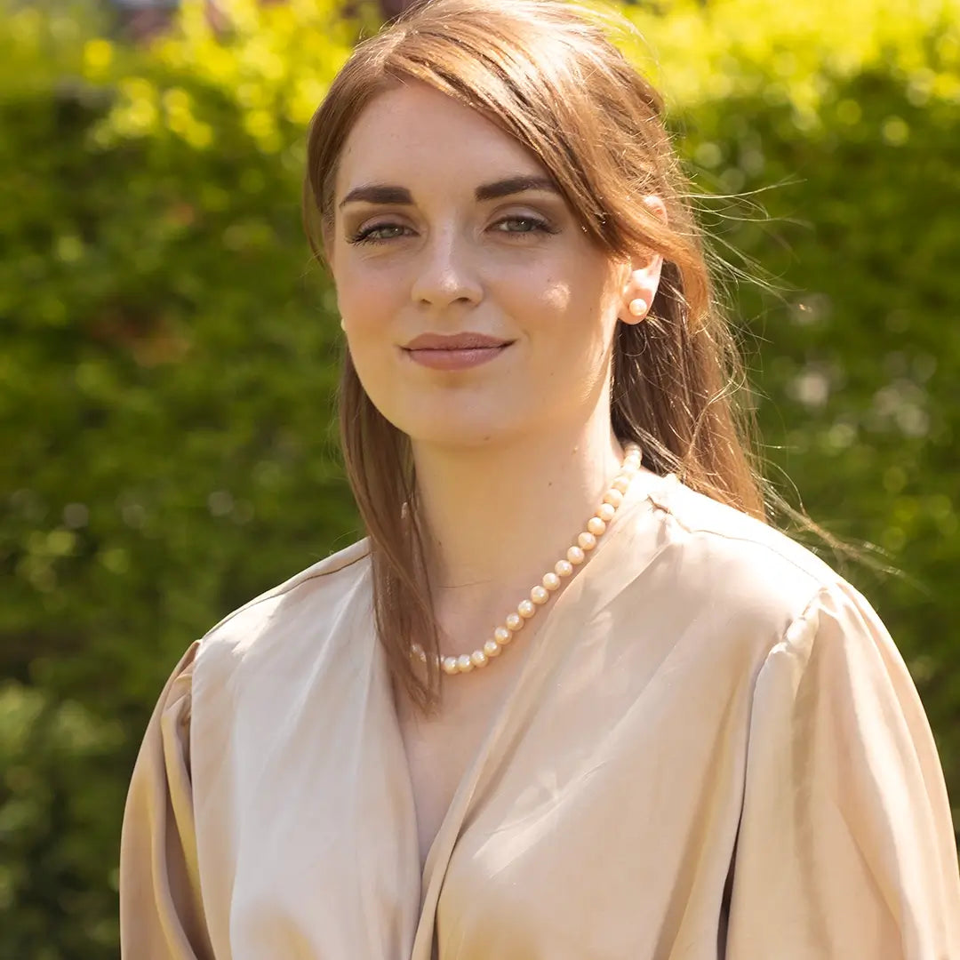 Woman wearing a champagne pearl necklace and matching pearl stud earrings, styled with a soft beige blouse, photographed outdoors in natural sunlight with greenery in the background.