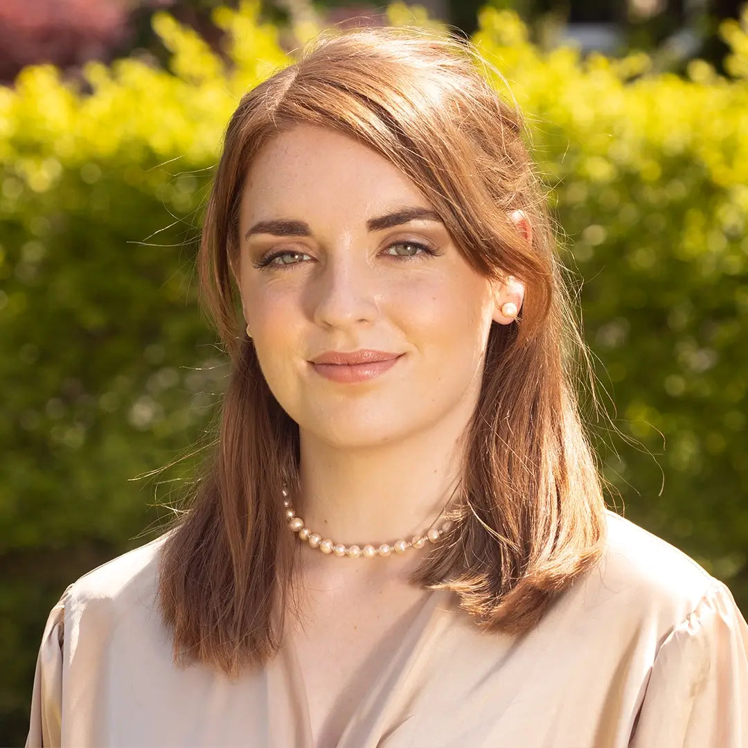 Woman wearing a delicate champagne pearl necklace and matching pearl stud earrings, styled with a soft beige blouse and photographed outdoors in warm natural sunlight with green foliage in the background.
