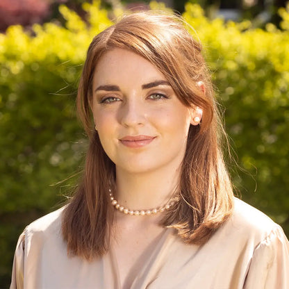 Woman wearing a delicate champagne pearl necklace and matching pearl stud earrings, styled with a soft beige blouse and photographed outdoors in warm natural sunlight with green foliage in the background.