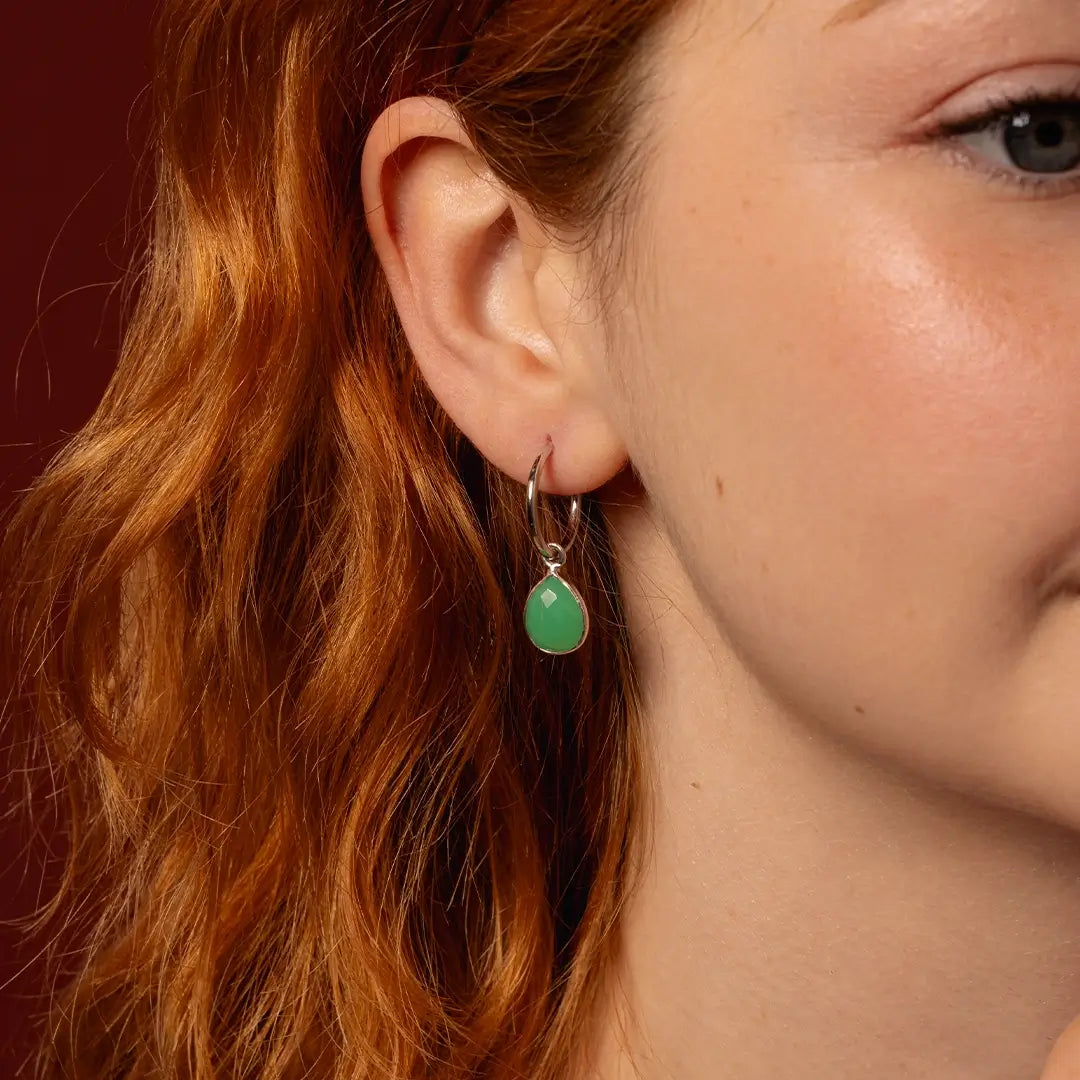 A close-up of a person with red hair wearing a silver hoop earring with a chrysoprase teardrop stone against a dark reddish-brown background.