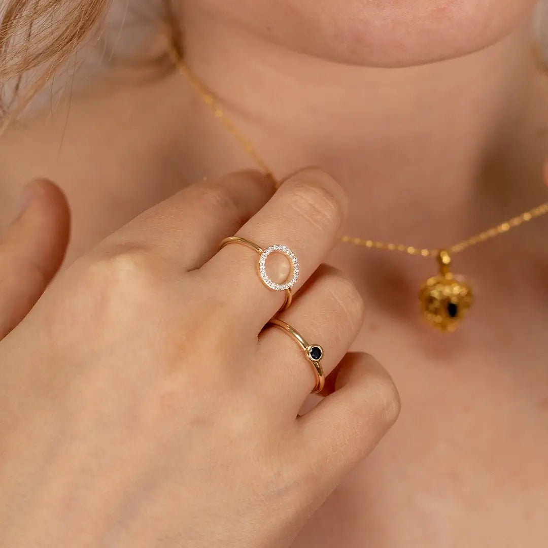 Close-up of a hand wearing a 9ct solid gold open circle diamond ring paired with a slim sapphire ring, alongside a fine gold necklace with a gemstone pendant in soft natural light.