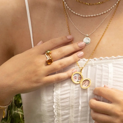 Gold photo locket necklace open with two images inside, styled with layered gold and silver necklaces and gemstone ring