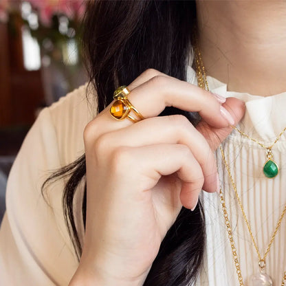 Gold ring with faceted amber gemstone worn on hand, paired with layered gold necklaces and green pendant