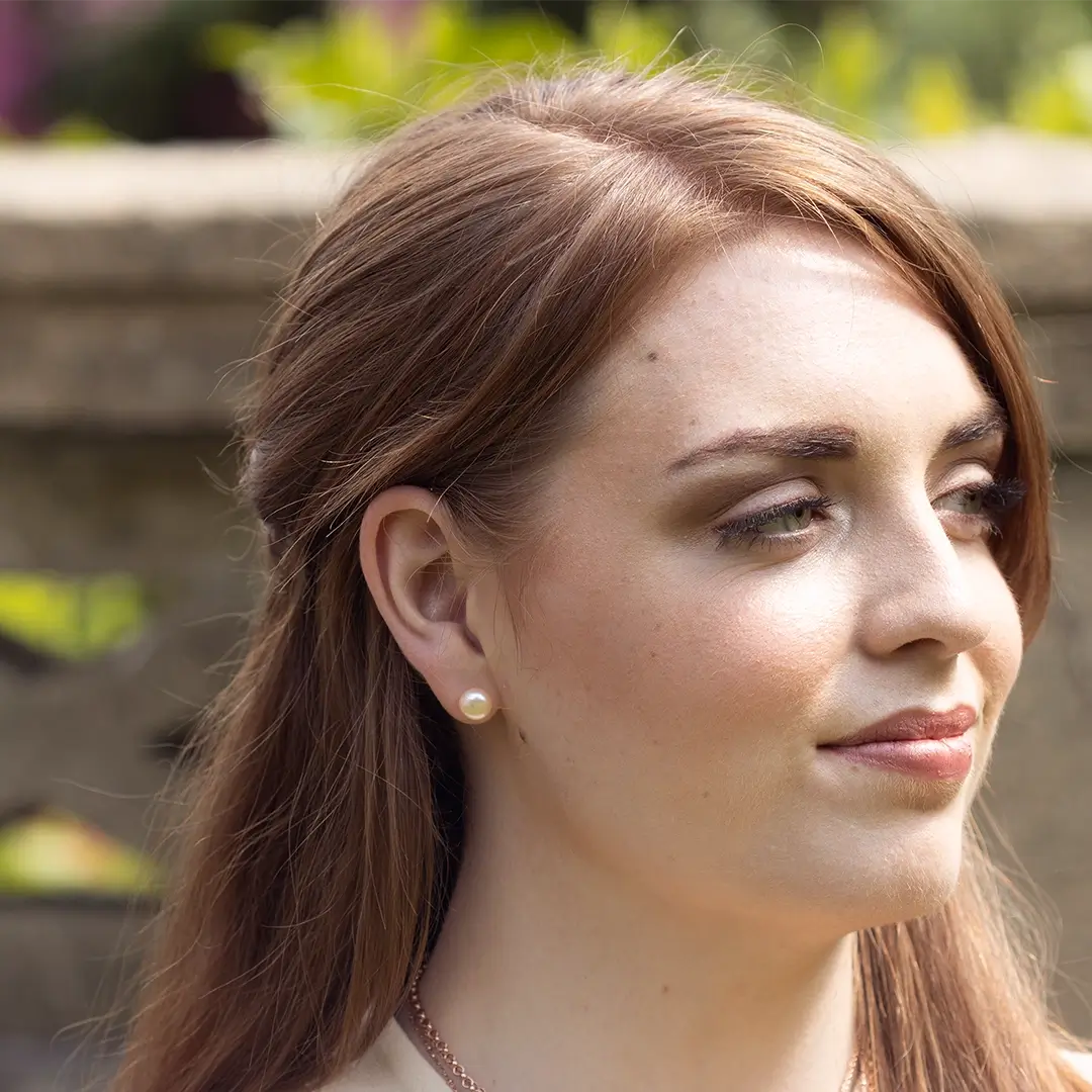 Woman wearing classic round pearl stud earrings, photographed outdoors in soft natural light with a blurred garden background.
