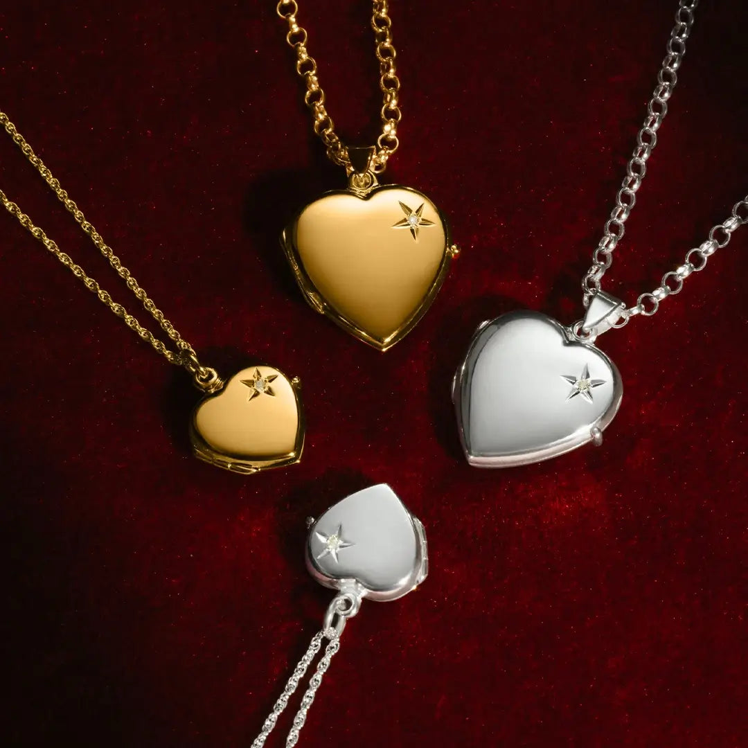 Four heart-shaped lockets displayed on a deep red velvet background. Two are gold and two are silver, each featuring a small diamond on the front. The lockets vary in size and hang from matching gold or silver chains.