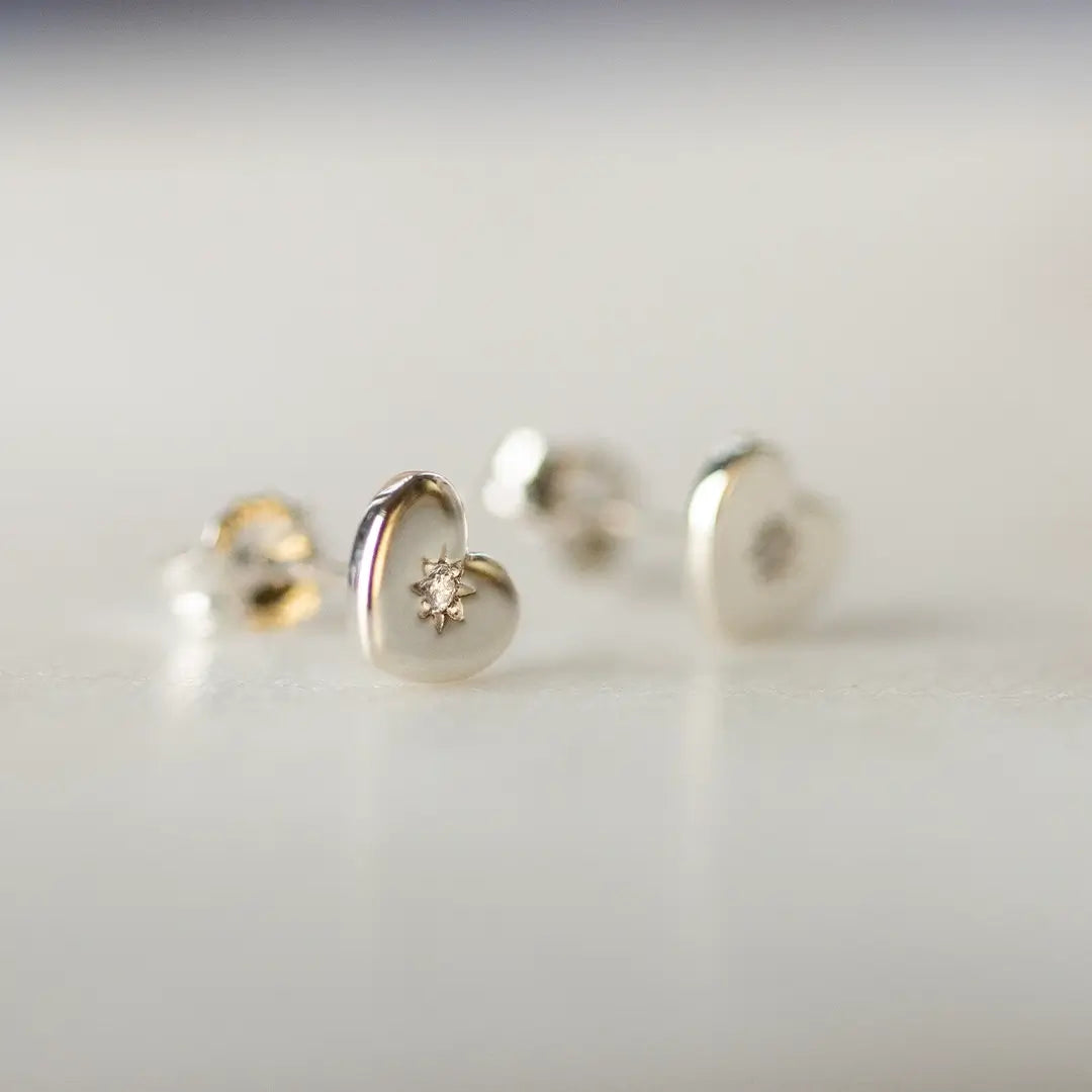 Sterling silver heart stud earrings with small diamond centre detail, featuring a polished finish and delicate starburst setting on a soft neutral background.
