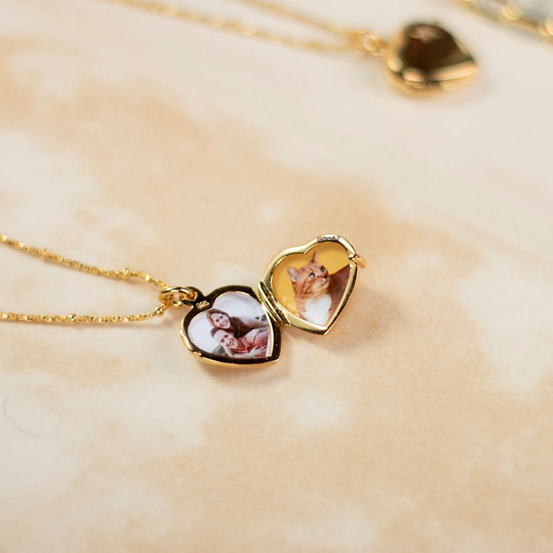 Lifestyle close-up of an open gold heart-shaped locket necklace, featuring two custom photos inside – one of a pet cat and one of a smiling couple. Set against a warm marbled background
