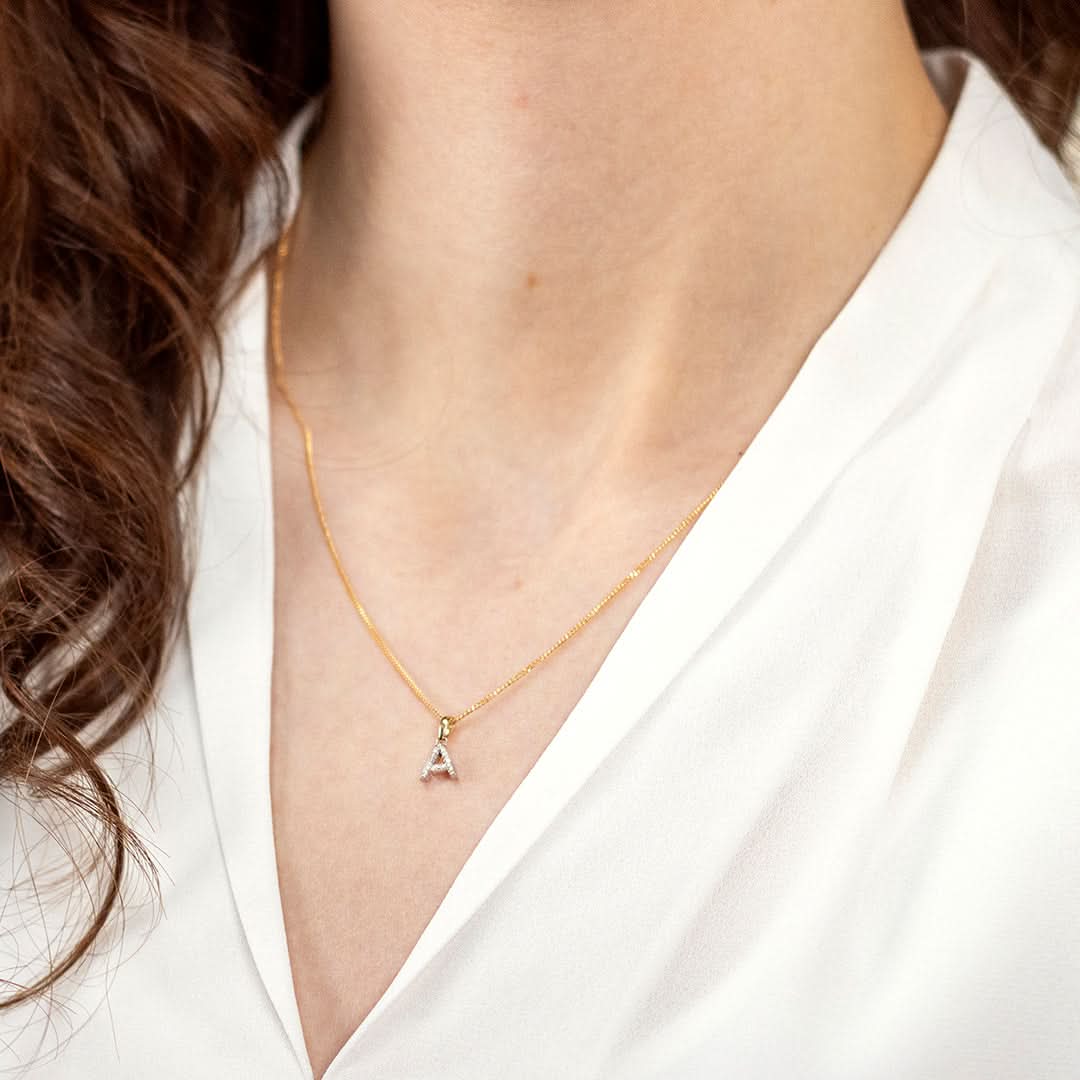 model wearing an "A" diamond initial necklace on a yellow gold curb chain. She is wearing a white v neck shit with her dark hair draped to one side.