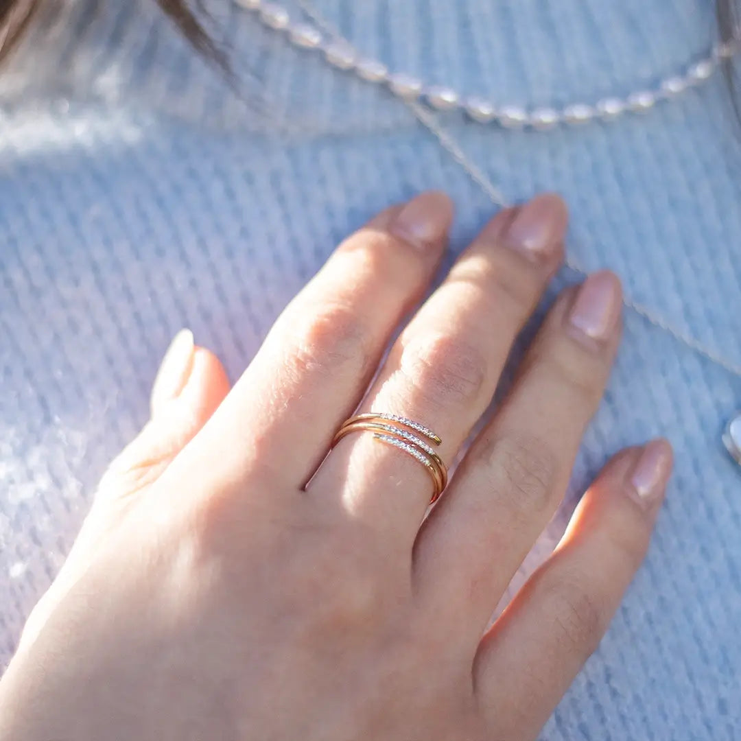 Close-up of a hand wearing a 9ct solid gold spiral ring with three wraparound bands, including two sparkling pavé diamond bands, styled with a fine silver beaded necklace in natural sunlight.