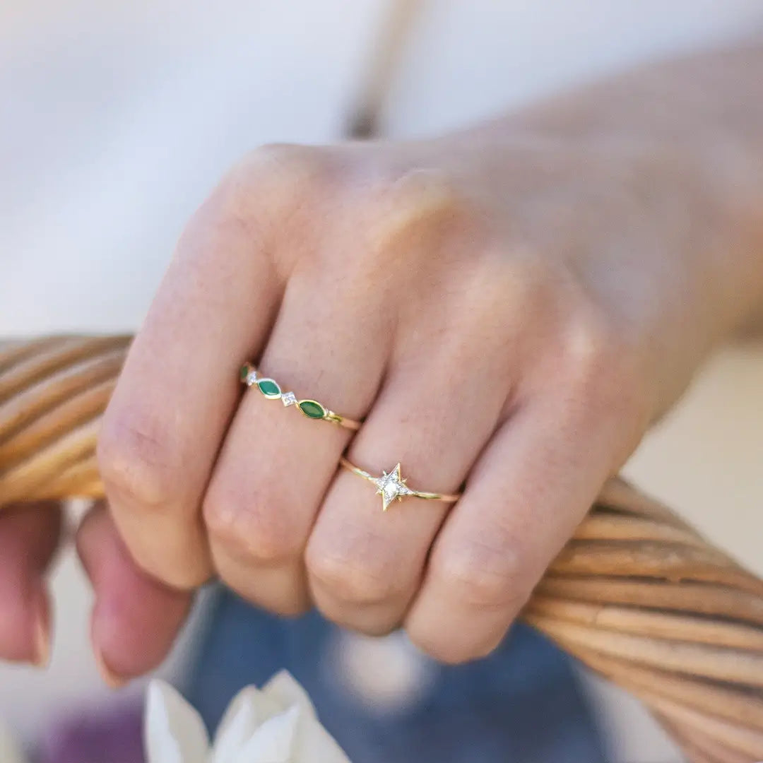 Model wearing a delicate 18ct gold vermeil diamond star ring alongside a green marquise gemstone band, with the hand resting on a woven rattan chair in soft natural light.