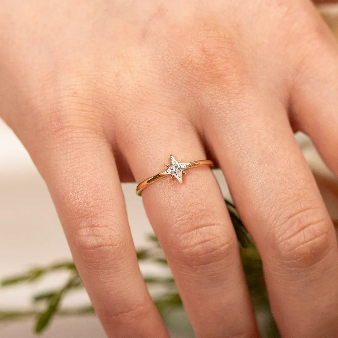 Close-up of a hand wearing a delicate 18ct gold vermeil diamond star ring, featuring a sparkling star-shaped setting on a fine polished band against a softly blurred floral background.