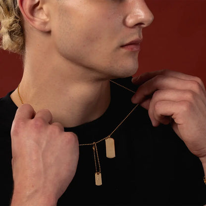 Close-up of male model wearing a gold double dog tag necklace, showing a blank tag and engraved initial tag