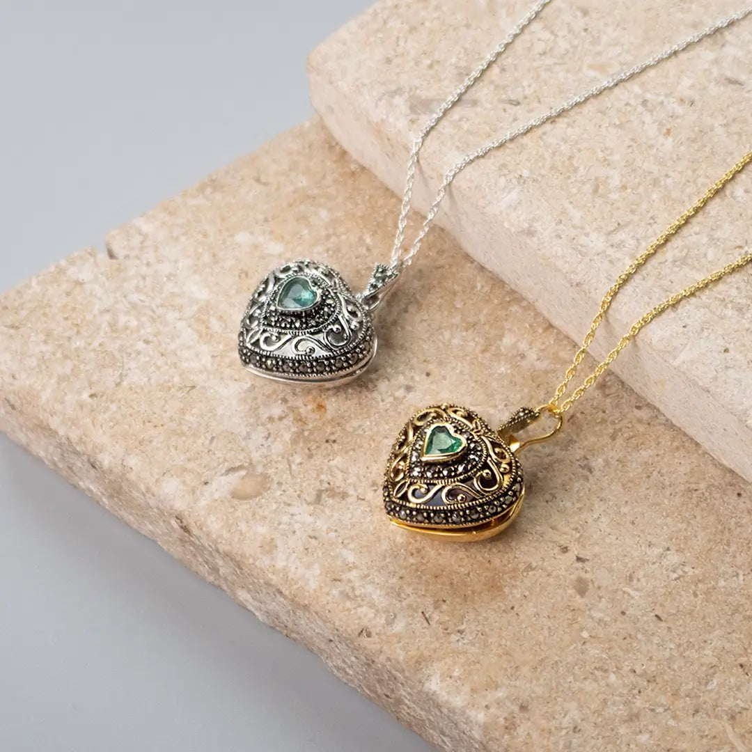 Two heart-shaped lockets with ornate vintage designs, one in silver and one in gold, both featuring small green heart-shaped gemstones in the center, placed on beige stone tiles.