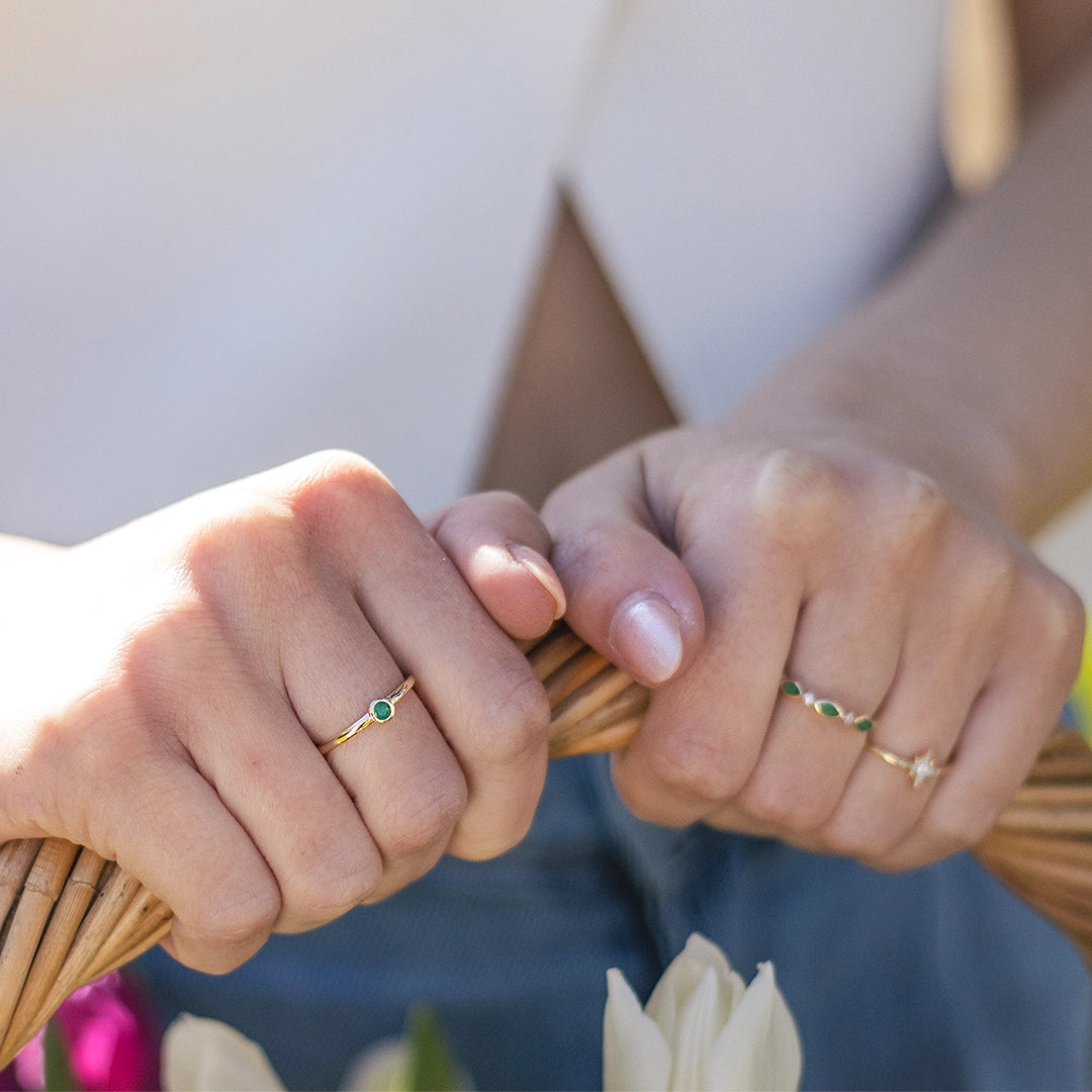 Solid gold emerald ring worn on the hand, featuring a vivid green emerald gemstone set on a fine gold band, styled alongside delicate gold stacking rings.