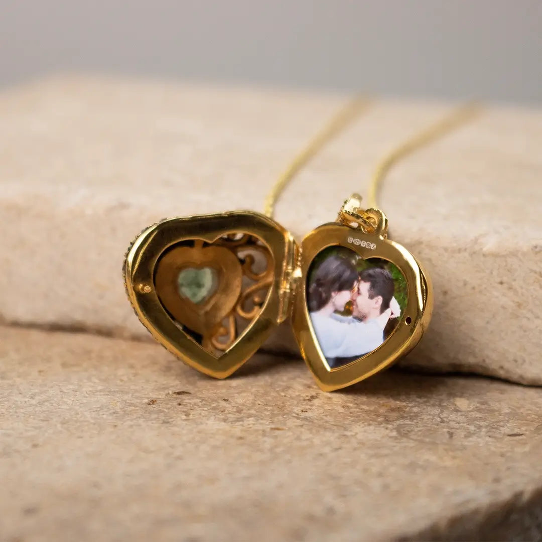 A gold heart-shaped locket opened to reveal a small green gemstone on one side and a photo of a couple embracing on the other, resting on a beige stone surface.