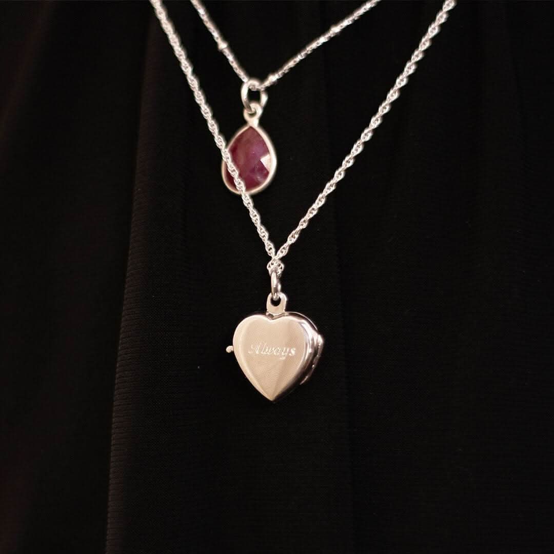 Lifestyle photo of a woman wearing a silver heart-shaped locket necklace engraved with the word “Always.” The locket is paired with a teardrop-shaped red gemstone pendant on a matching chain. Styled over a black outfit