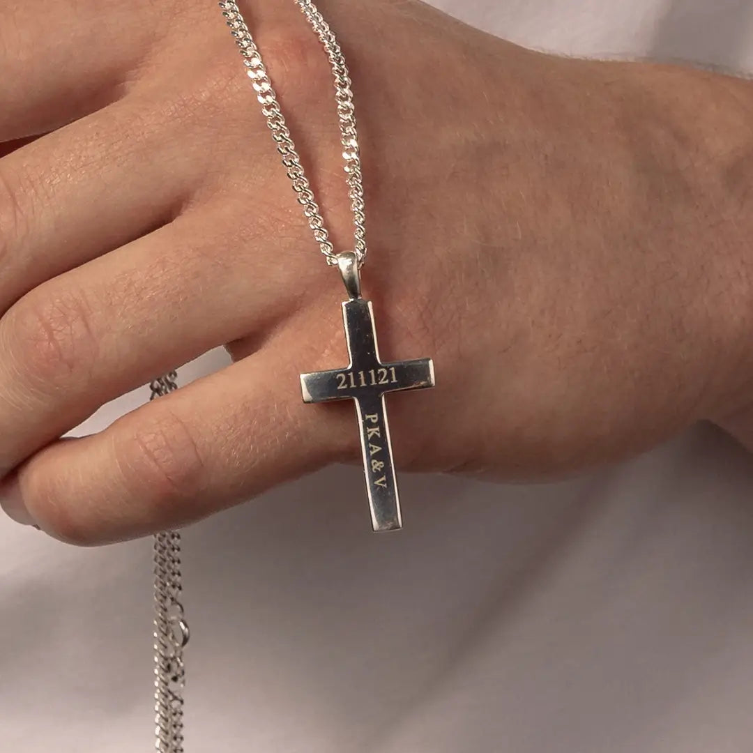 A close-up of a hand wearing a silver cross pendant necklace engraved with '211121' and 'PAXABY'.