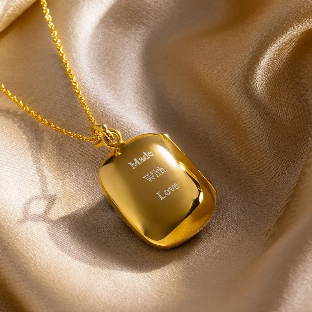Gold rectangular locket necklace engraved with “Made With Love” on a polished finish, styled on champagne satin fabric.