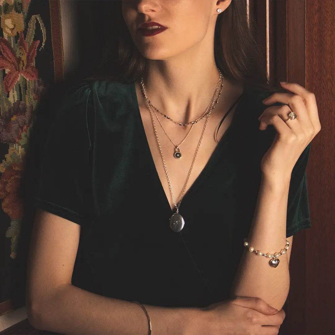 Woman wearing a sterling silver oval locket necklace layered with fine silver chains, styled with a deep green velvet dress in warm, vintage lighting.