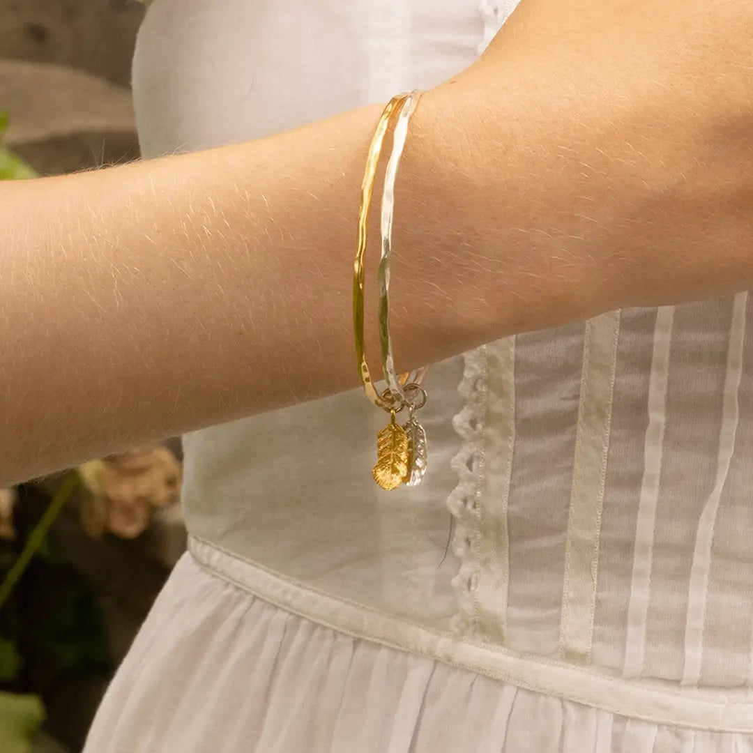 Hammered sterling silver and gold bangle bracelet worn on the wrist, featuring delicate feather charms in mixed metal tones.
