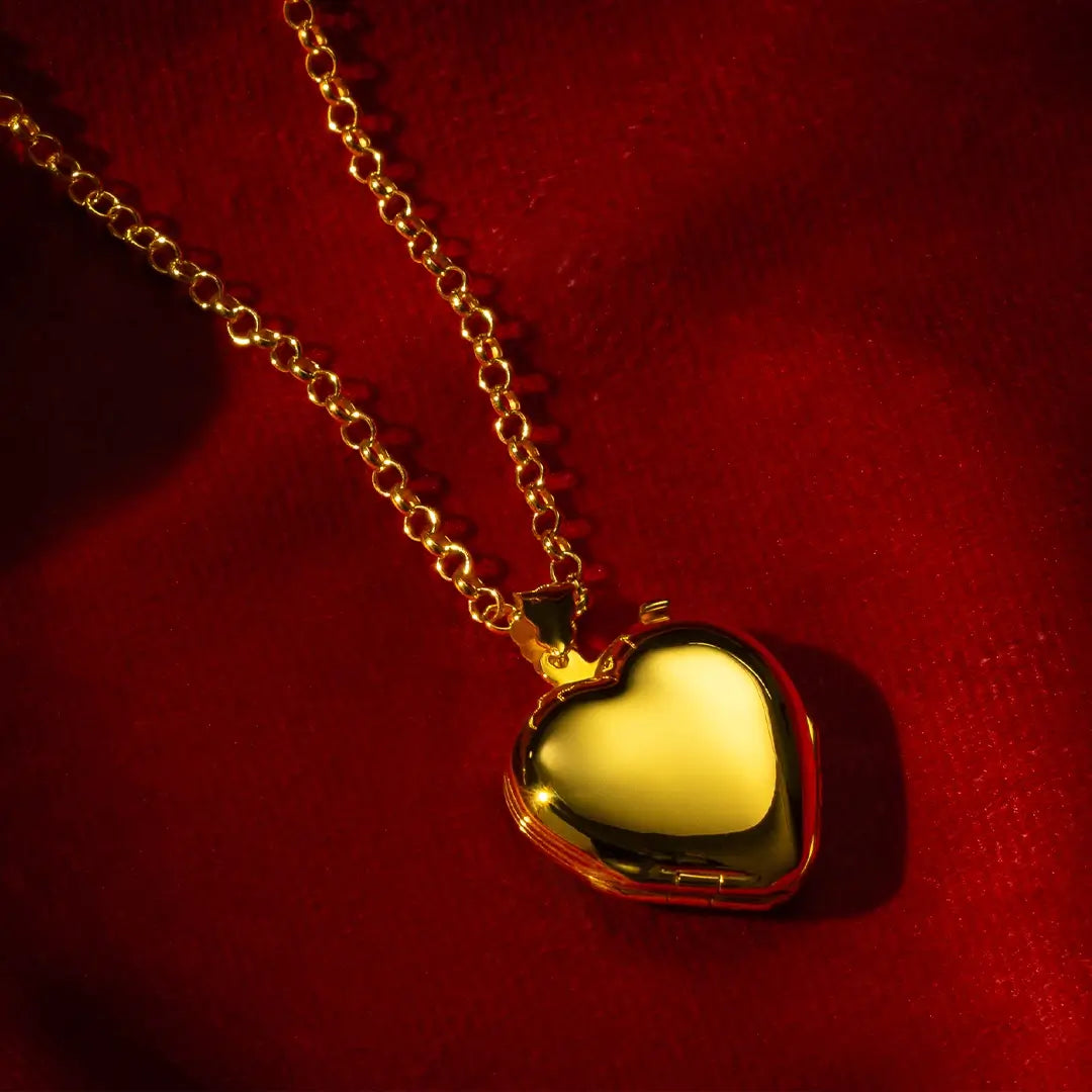 Polished gold heart locket necklace with a chunky gold chain, displayed against rich red fabric in dramatic lighting.