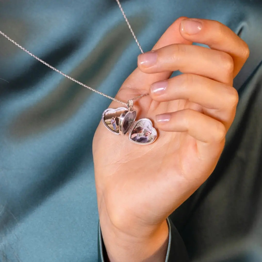 Model holding an open sterling silver heart locket necklace showing personalised photos inside, worn with a satin teal top and delicate silver chain.
