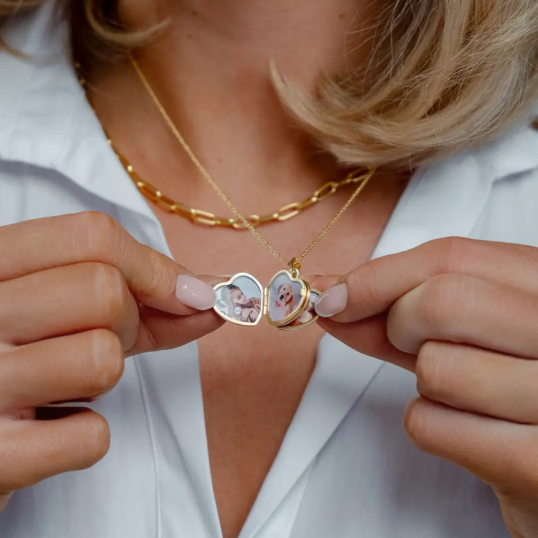 Woman wearing a white shirt and layered gold necklaces, opening a gold heart-shaped photo locket to reveal four personalised photographs inside. The locket hangs from a fine gold chain and is held open with both hands, highlighting its four-photo design and polished finish.