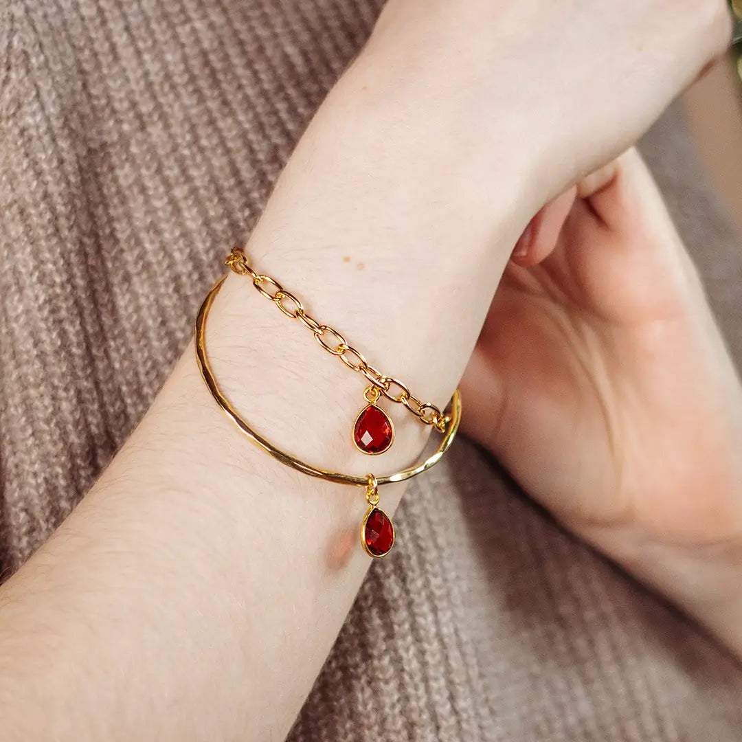 Gold bangle bracelet with faceted garnet teardrop charm and delicate chain detail, worn on wrist against a soft knit background
