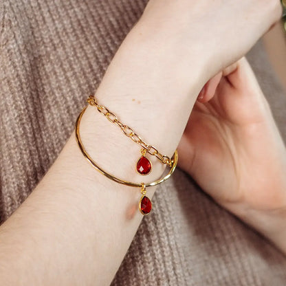 Gold bangle bracelet with faceted garnet teardrop charm and delicate chain detail, worn on wrist against a soft knit background