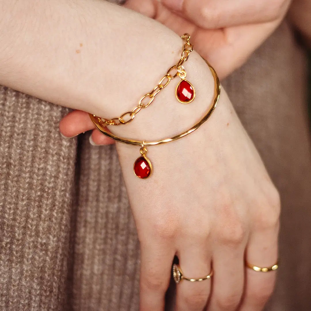 Close-up of gold hammered bangle with dangling garnet gemstone charm and fine chain, styled on wrist with gold rings