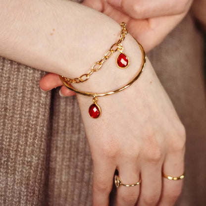 Close-up of gold hammered bangle with dangling garnet gemstone charm and fine chain, styled on wrist with gold rings