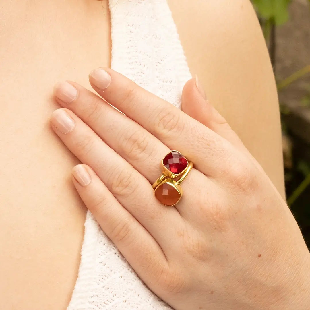 Gold statement stacked rings with two faceted gemstones—deep red garnet and warm amber carnelian—worn on a hand against a soft, neutral background.