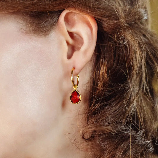 Close-up of gold vermeil hoop earrings with oval-cut garnet gemstone drop, worn on ear, showcasing deep red January birthstone.