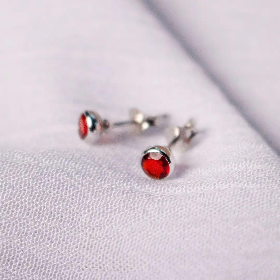 Sterling silver stud earrings with round-cut garnet gemstones in a polished bezel setting, featuring deep red January birthstones on a soft white fabric background.