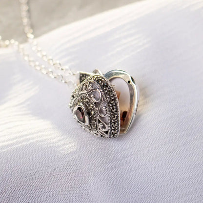 Side view of an open vintage-style sterling silver heart locket with decorative metalwork and garnet stone, resting on white fabric.