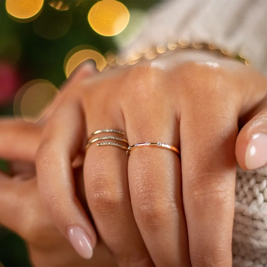 Close-up of a hand wearing a delicate 9ct solid gold diamond ring with five bezel-set diamonds, styled alongside two slim diamond stacking rings, with warm festive lights softly blurred in the background.
