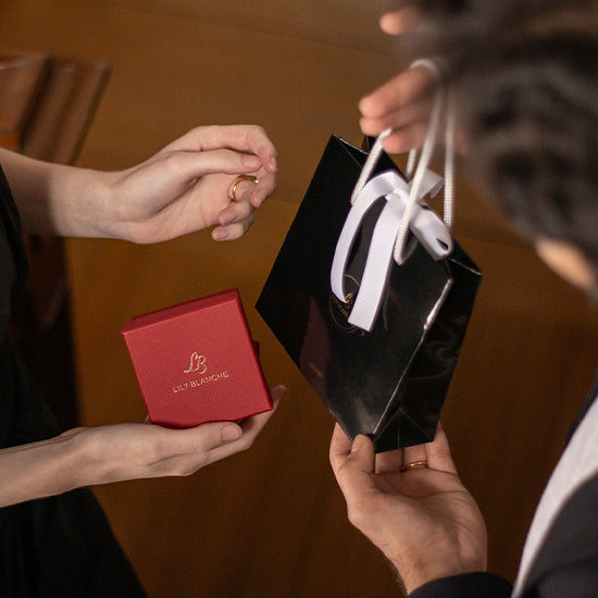 Close up image of a man and woman's hands exchanging gifts. She is holding a red Lily Blanche gift box and he is holding up a black and white ribbon-tied Lily Blanche gift bag. 