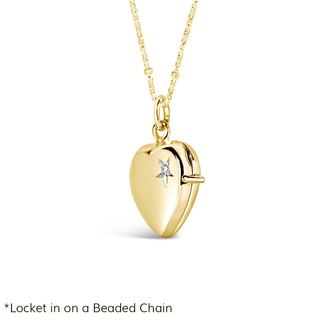 product image of a gold heart-shaped locket necklace featuring a delicate diamond-set starburst engraving. The locket hangs on a fine beaded gold chain and is photographed on a clean white background