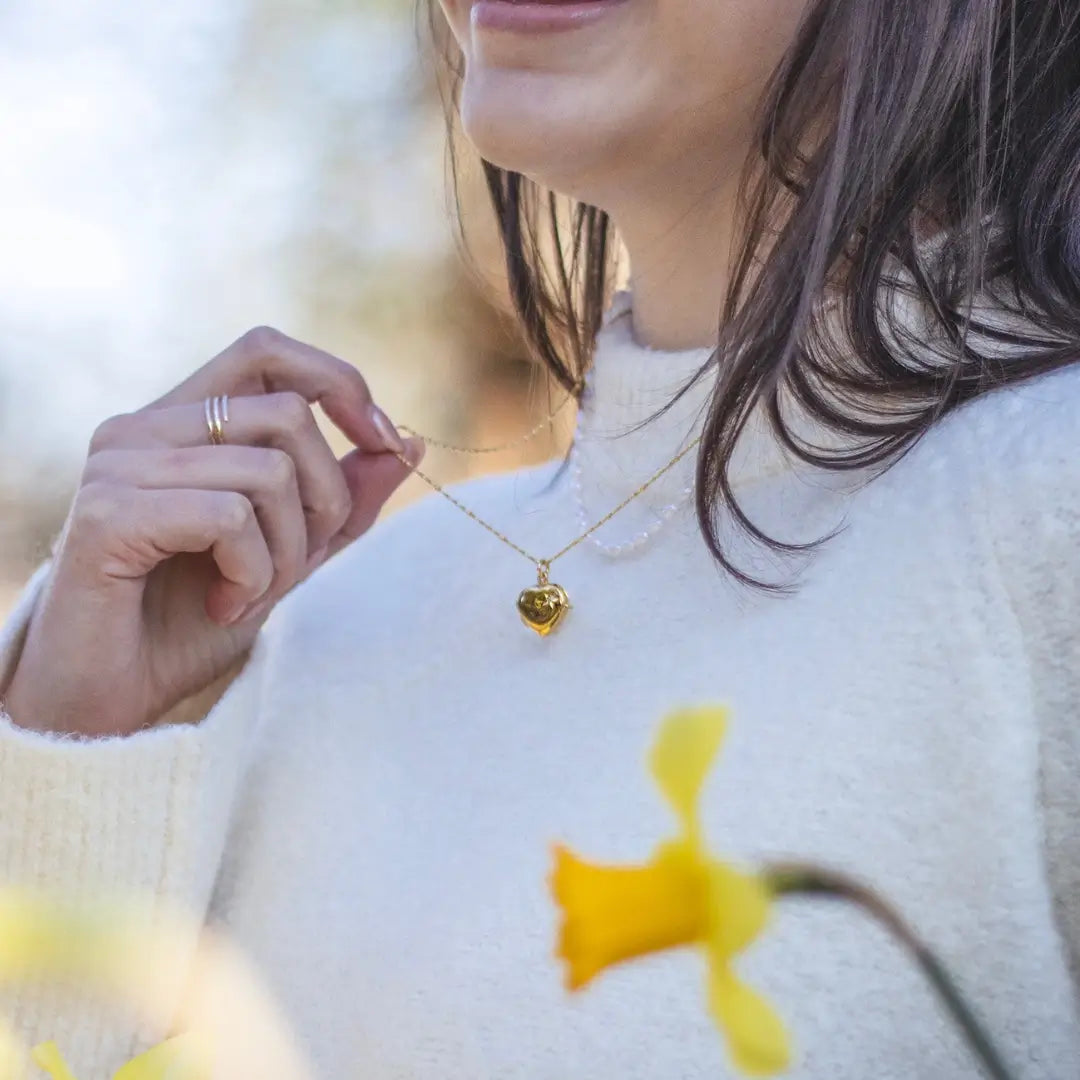 A person smiling while holding a gold heart-shaped locket necklace, surrounded by yellow flowers outdoors.