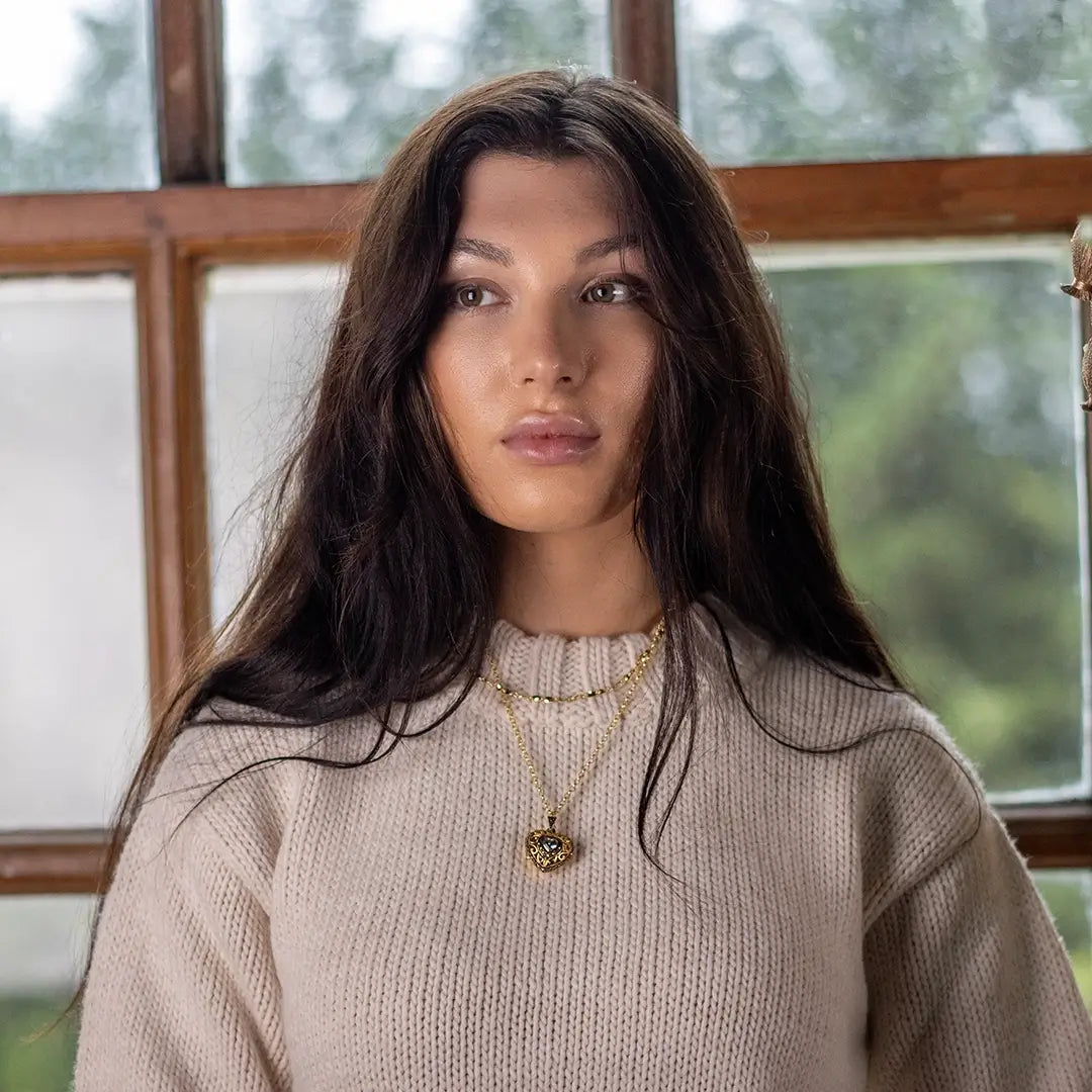 A person wearing a gold heart-shaped locket necklace with a blue gemstone centre and 16"gold chain, standing indoors by a window.