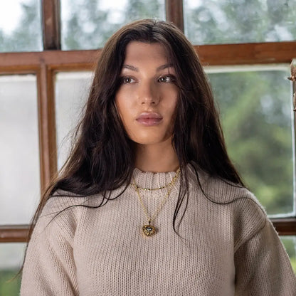 A person wearing a gold heart-shaped locket necklace with a blue gemstone centre and 16"gold chain, standing indoors by a window.