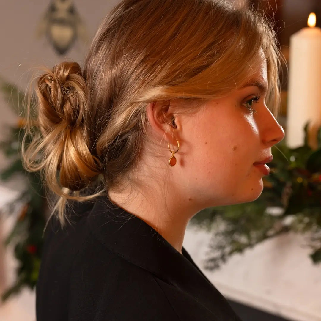 Side profile of a woman in a black blazer wearing gold hoop earrings with teardrop-shaped carnelian gemstones, in a softly lit indoor setting
